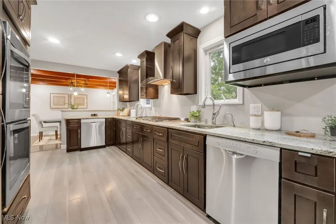 Kitchen featuring stainless steel appliances, a sink, dark brown cabinets, light wood-style floors, and light stone counters