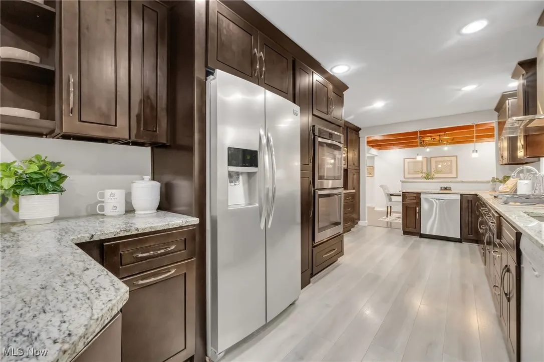 Kitchen featuring appliances with stainless steel finishes, dark brown cabinetry, light wood-style floors, light stone counters, and open shelves