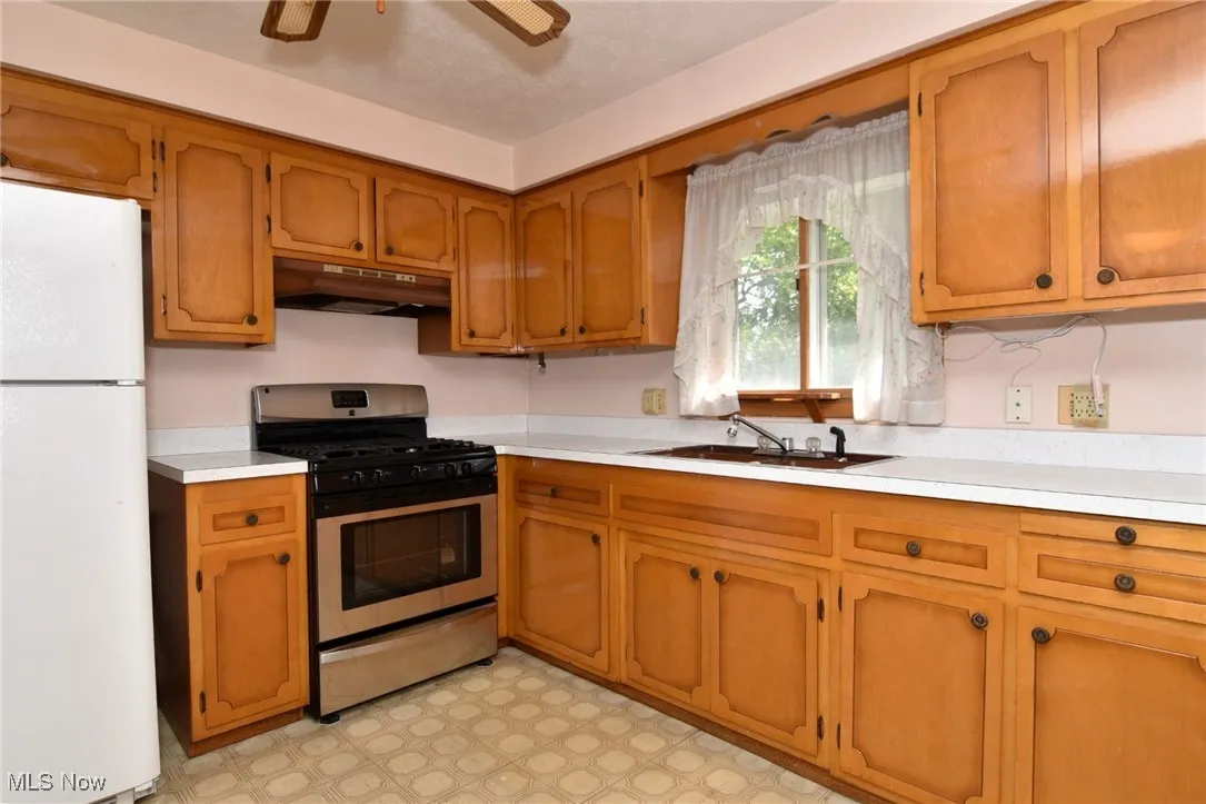 Kitchen featuring freestanding refrigerator, under cabinet range hood, gas range, light countertops, and brown cabinets
