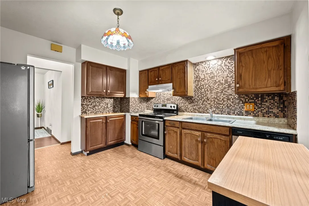 Kitchen featuring appliances with stainless steel finishes, a sink, under cabinet range hood, backsplash, and pendant lighting