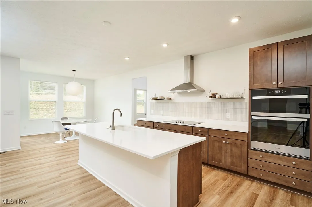 Kitchen with wall chimney range hood, light wood-style flooring, black electric stovetop, a sink, and multiple ovens