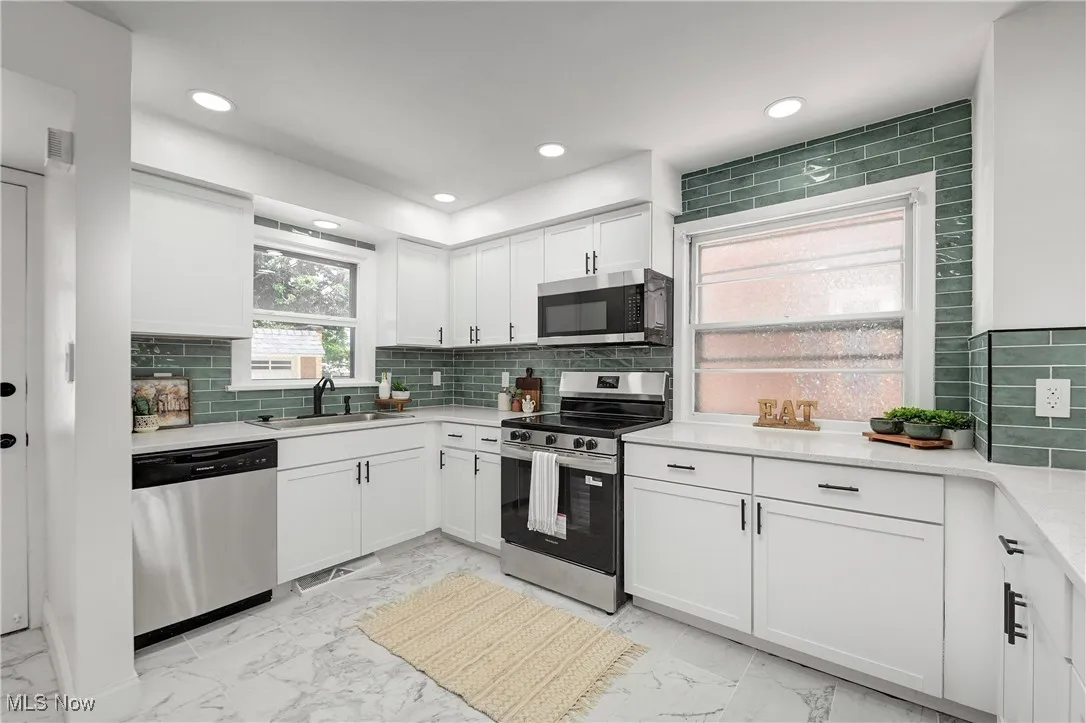 Kitchen with stainless steel appliances, light marble finish flooring, white cabinetry, light countertops, and backsplash.