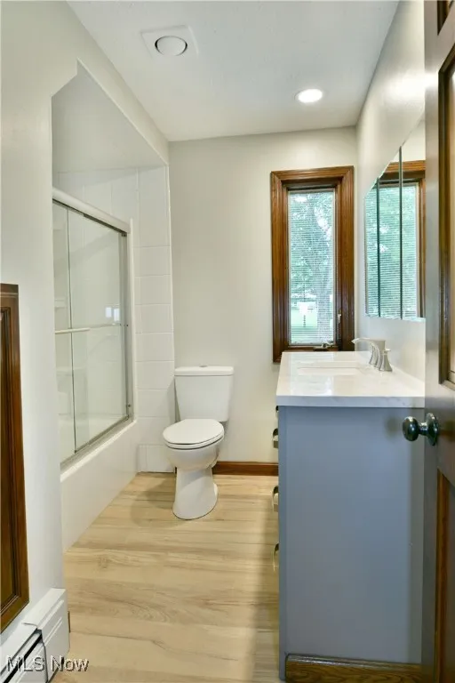 Bathroom with baseboard heating, toilet, vanity, wood finished floors, and bath / shower combo with glass door