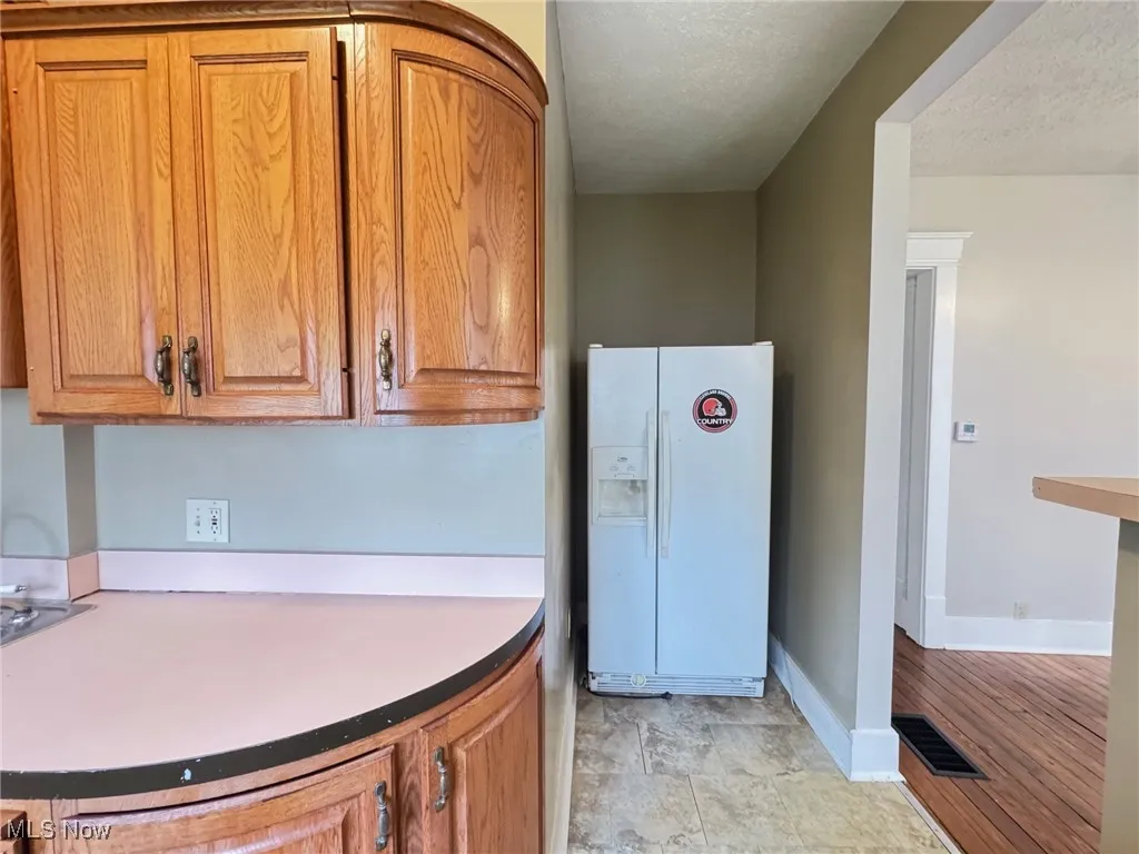 First floor Kitchen with white fridge with ice dispenser, brown cabinetry, baseboards, and a textured ceiling