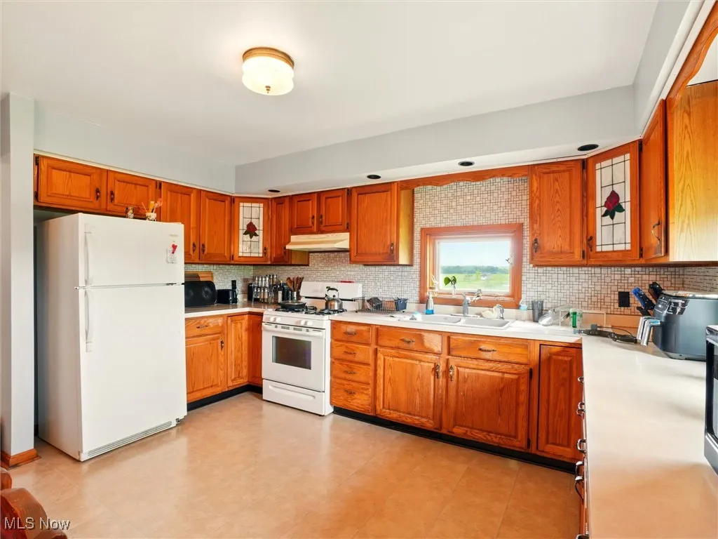 Kitchen featuring white appliances, a sink, under cabinet range hood, light countertops, and brown cabinetry