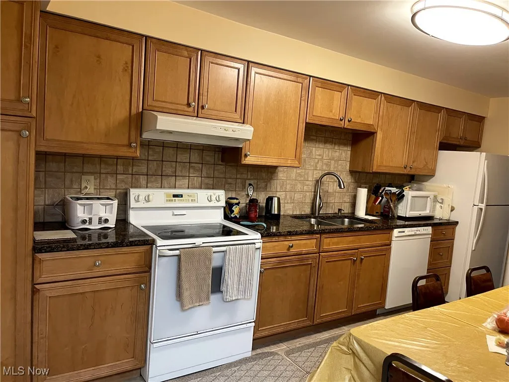 Kitchen featuring under cabinet range hood, white appliances, a sink, brown cabinetry, and decorative backsplash