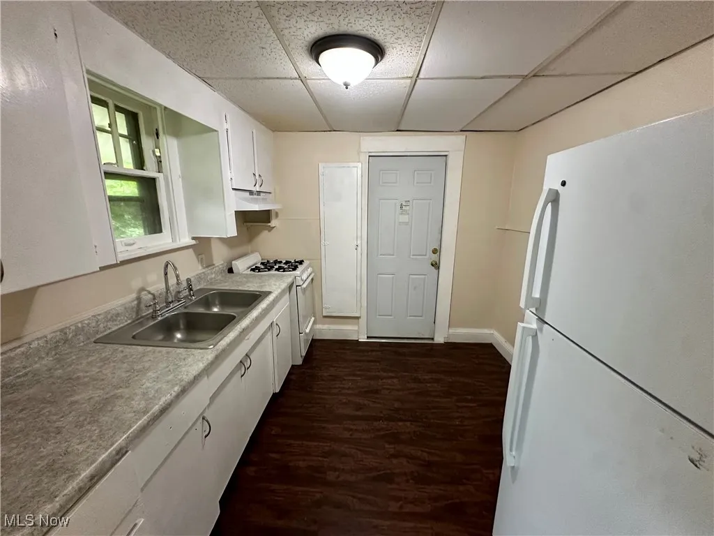 Kitchen with white appliances, a sink, under cabinet range hood, a paneled ceiling, and white cabinetry