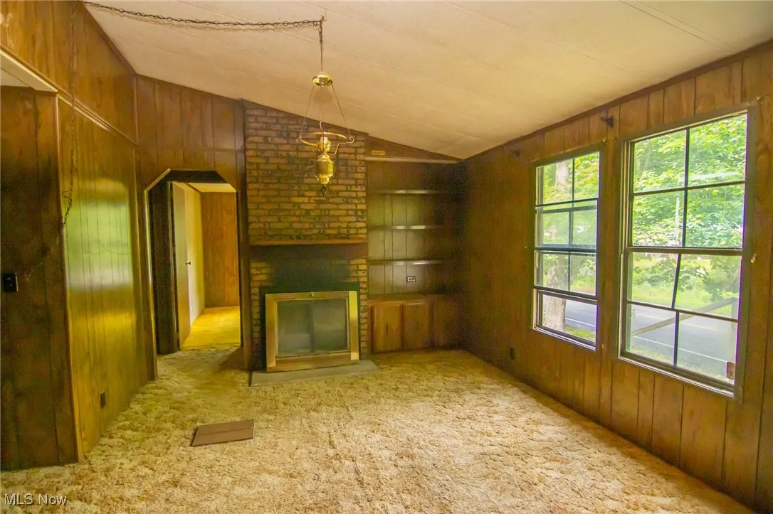 Unfurnished living room with wooden walls, lofted ceiling, a brick fireplace, and carpet