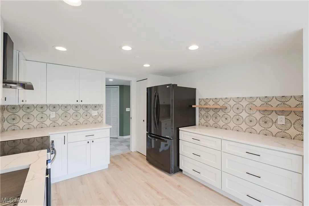 Kitchen featuring freestanding refrigerator, wall chimney exhaust hood, light stone counters, white cabinetry, and decorative backsplash