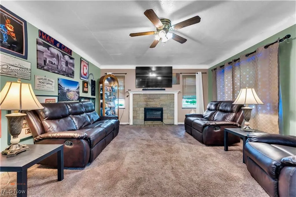 Living room with carpet floors, a ceiling fan, a stone fireplace, and baseboards