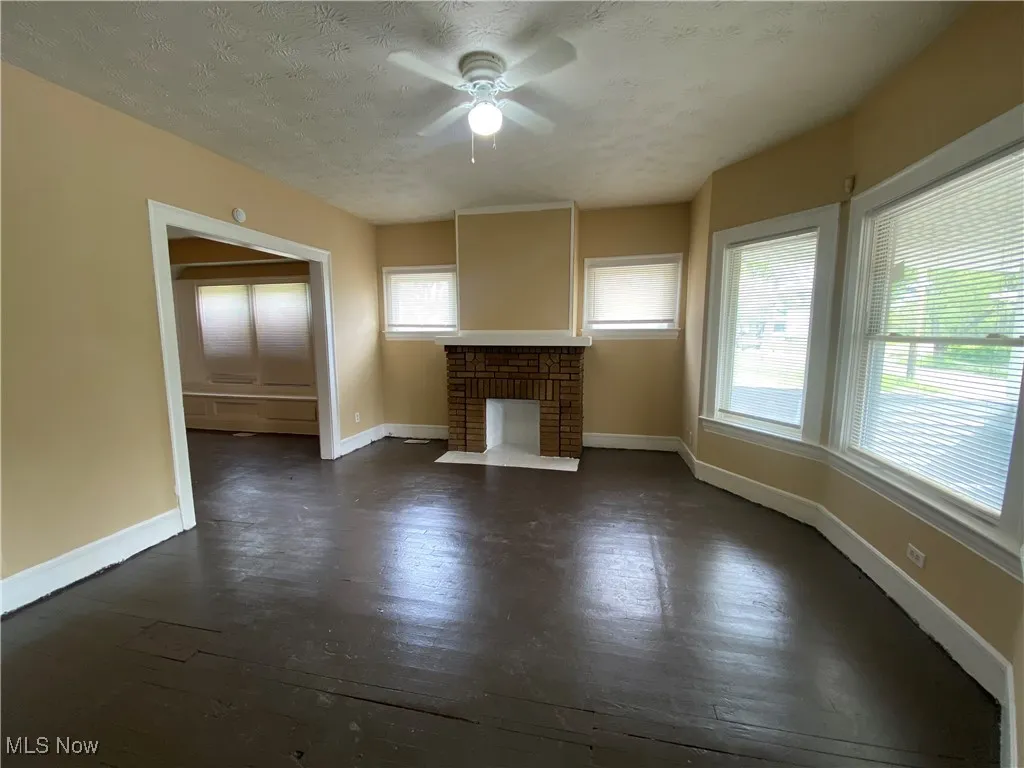 Unfurnished living room with a ceiling fan, baseboards, a brick fireplace, and dark wood-style floors