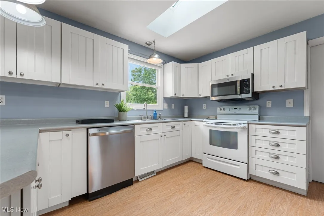 Kitchen featuring stainless steel appliances, a skylight, light wood-type flooring, light countertops, and white cabinetry