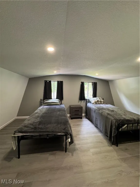 Bedroom featuring light wood-type flooring, a textured ceiling, vaulted ceiling, baseboards, and multiple windows