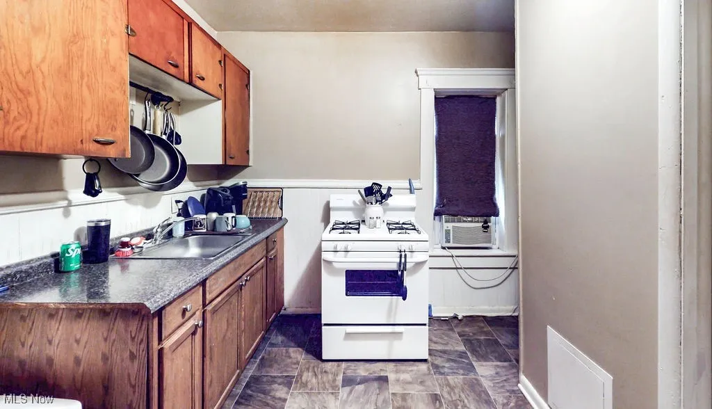 Kitchen with white gas range oven, cooling unit, dark countertops, a sink, and brown cabinets