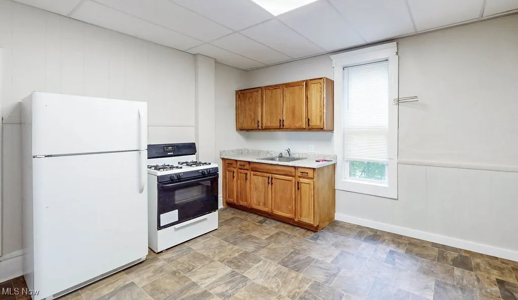 Kitchen with white appliances, a sink, a paneled ceiling, light countertops, and brown cabinetry