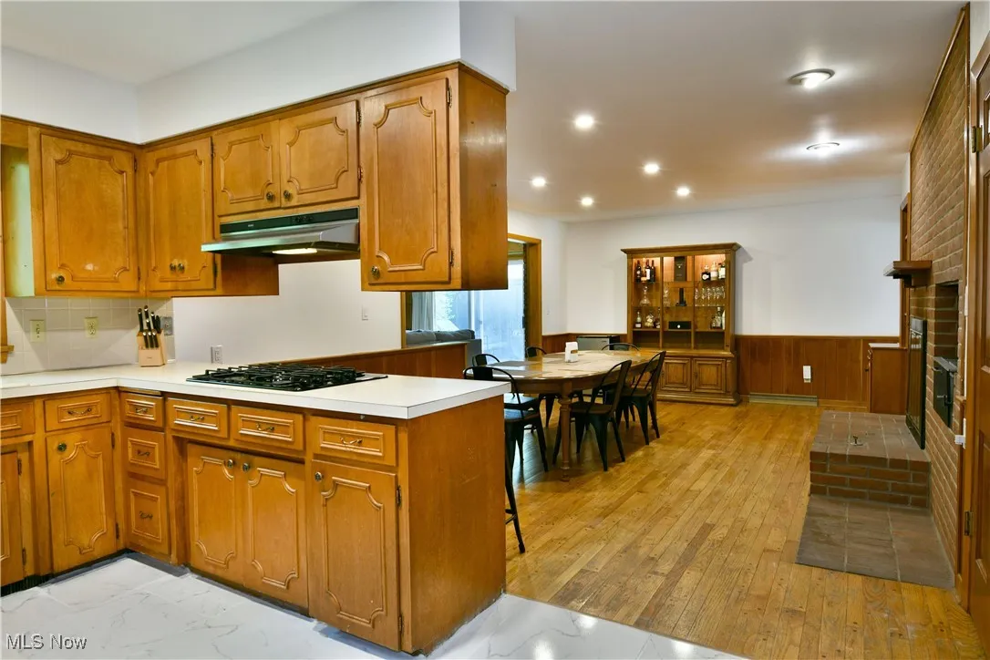 Kitchen featuring under cabinet range hood, black gas stovetop, brown cabinetry, a wainscoted wall, and light countertops