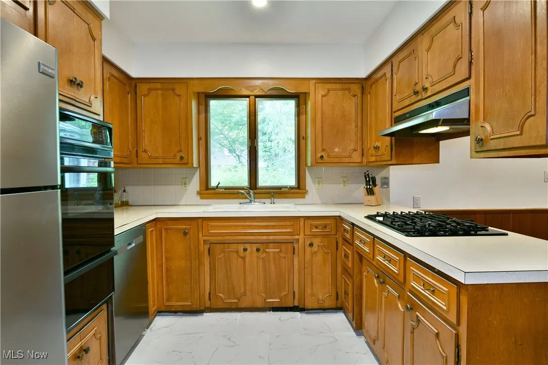 Kitchen featuring a sink, under cabinet range hood, black appliances, light marble finish floors, and brown cabinets