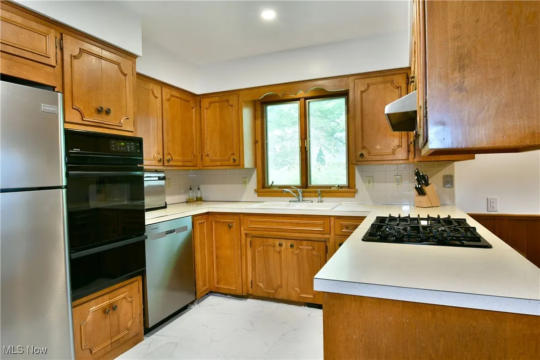 Kitchen featuring black appliances, a sink, under cabinet range hood, light marble finish flooring, and brown cabinets