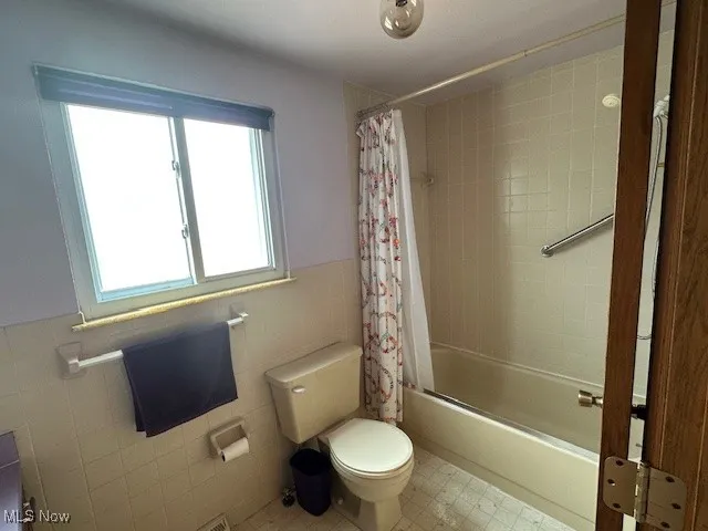 Full bathroom featuring tile walls, toilet, shower / bath combination with curtain, a wainscoted wall, and tile patterned floors