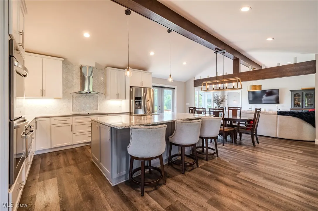 Kitchen featuring wall chimney range hood, appliances with stainless steel finishes, light stone countertops, a center island with sink, and backsplash