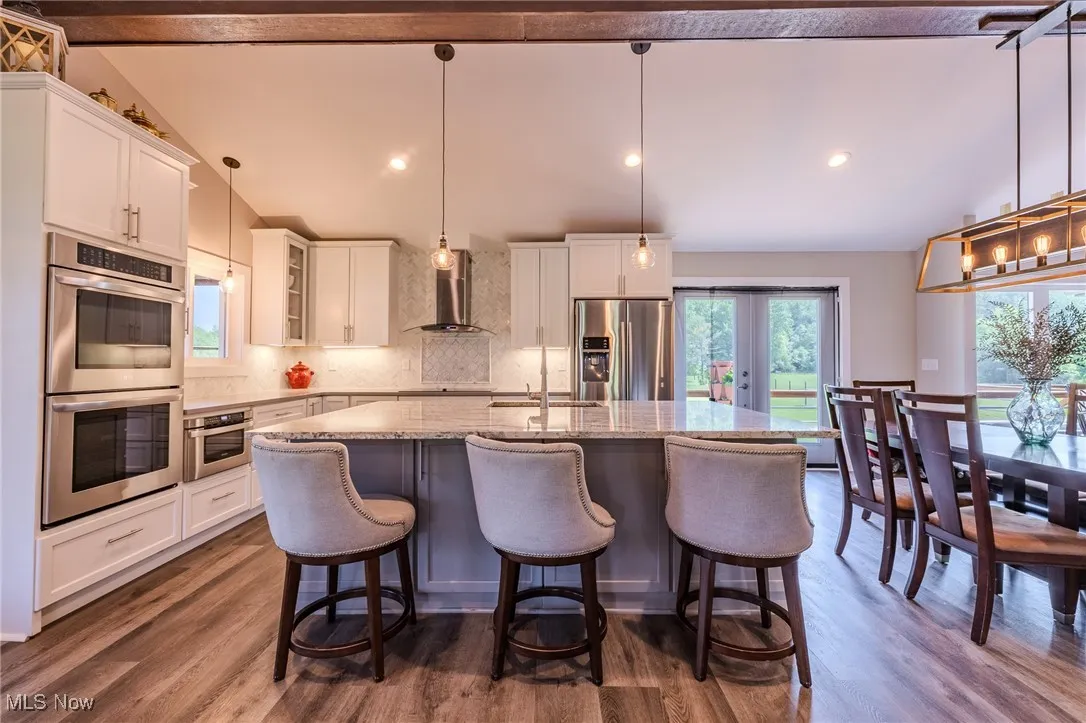 Kitchen with appliances with stainless steel finishes, wall chimney range hood, a kitchen island with sink, tasteful backsplash, and white cabinets
