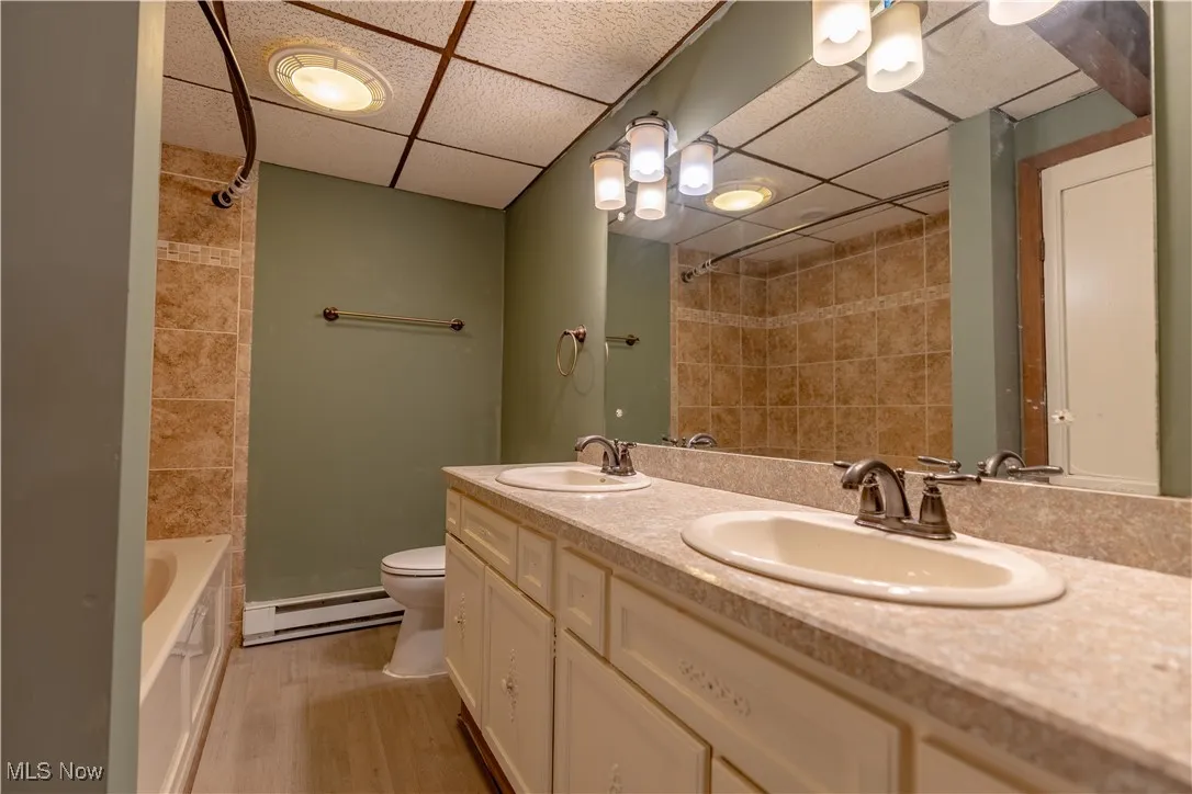 Bathroom featuring a baseboard heating unit, a paneled ceiling, toilet, double vanity, and wood finished floors