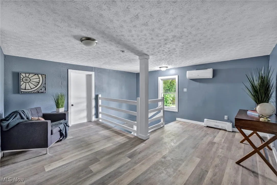second floor landing area with a wall mounted AC, wood finished floors, a baseboard heating unit, a textured ceiling, and baseboards