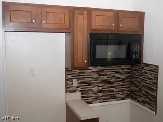 Kitchen featuring black microwave, decorative backsplash, brown cabinets, and light countertops