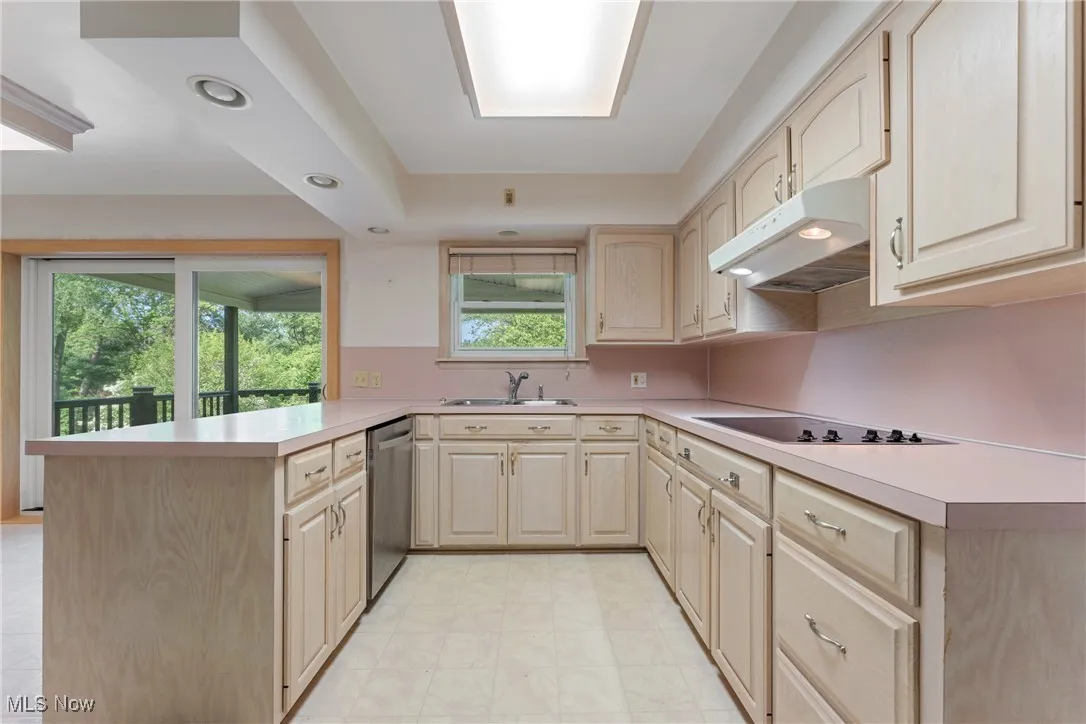 Kitchen featuring light brown cabinetry, stainless steel dishwasher, a peninsula, under cabinet range hood, and black electric stovetop