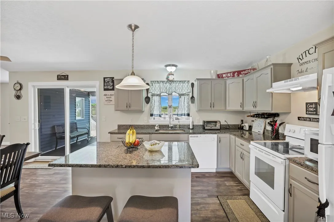 Kitchen featuring white appliances, a sink, under cabinet range hood, dark wood-style flooring, and a breakfast bar area