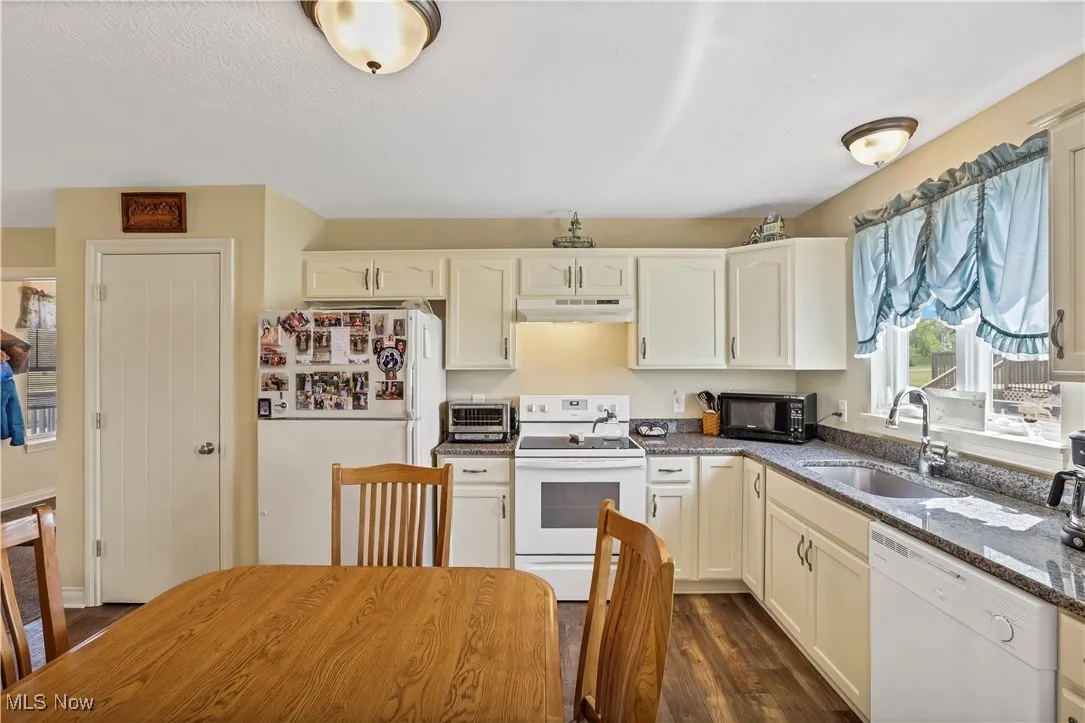 Kitchen with white appliances, under cabinet range hood, a sink, dark stone countertops, and dark wood-style flooring