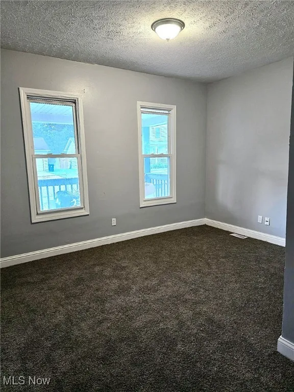 Empty room featuring dark colored carpet, plenty of natural light, a textured ceiling, and baseboards