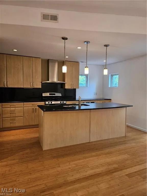 Kitchen with wall chimney range hood, light brown cabinets, stainless steel gas stove, dark countertops, and light wood finished floors