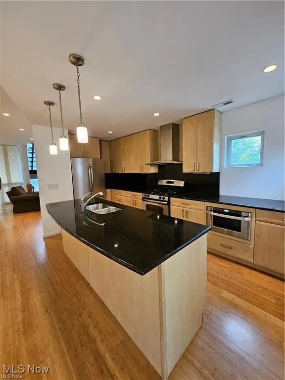 Kitchen with stainless steel appliances, wall chimney range hood, a sink, light wood-style flooring, and light brown cabinets