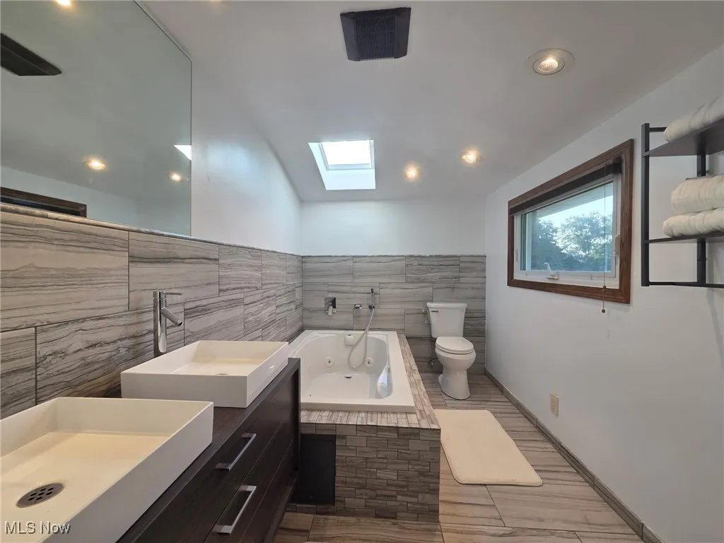 Full bathroom with tile walls, a skylight, vanity, a jetted tub, and vaulted ceiling