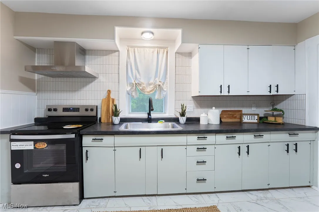 Kitchen with wall chimney exhaust hood, electric range, dark countertops, light marble finish floors, and tasteful backsplash