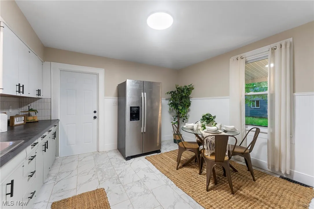 Kitchen with stainless steel fridge, dark countertops, light marble finish flooring, white cabinets, and wainscoting