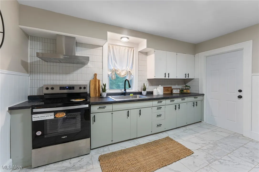 Kitchen featuring electric range, light marble finish floors, wall chimney exhaust hood, dark countertops, and a wainscoted wall