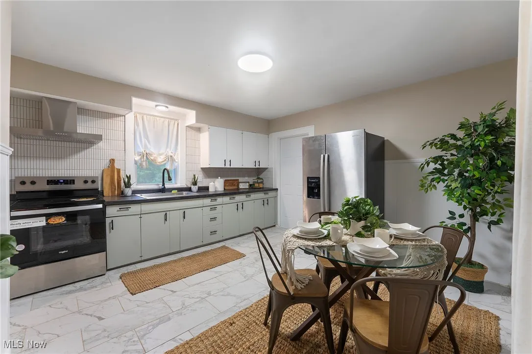 Kitchen featuring stainless steel appliances, light marble finish floors, wall chimney range hood, and dark countertops