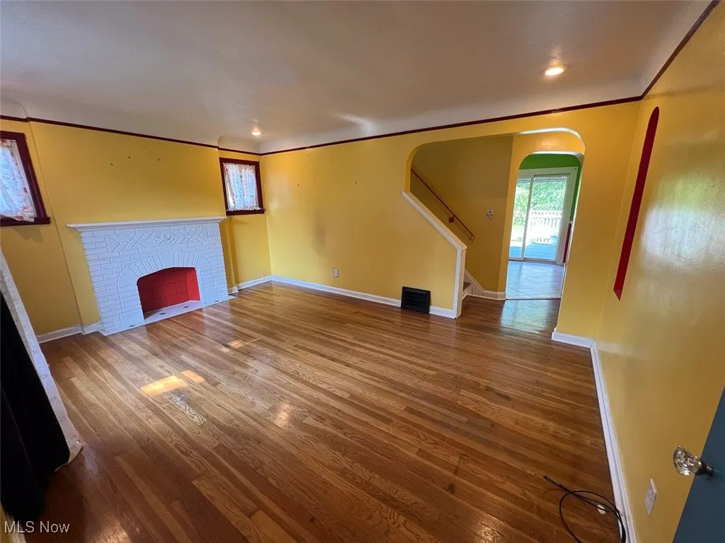 Unfurnished living room with arched walkways, wood finished floors, a brick fireplace, stairway, and crown molding