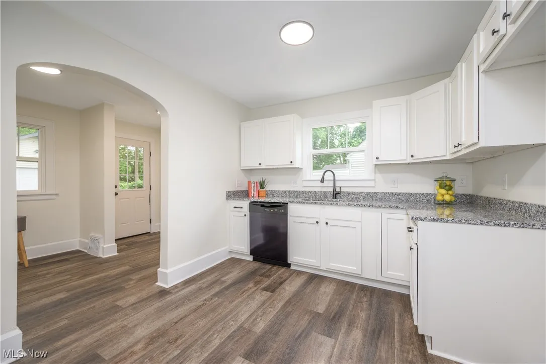 Kitchen with black dishwasher, light stone countertops, dark wood finished floors, white cabinets, and arched walkways
