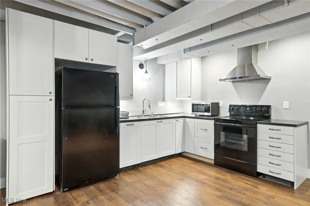 Kitchen with black appliances, wall chimney exhaust hood, dark countertops, dark wood finished floors, and white cabinets