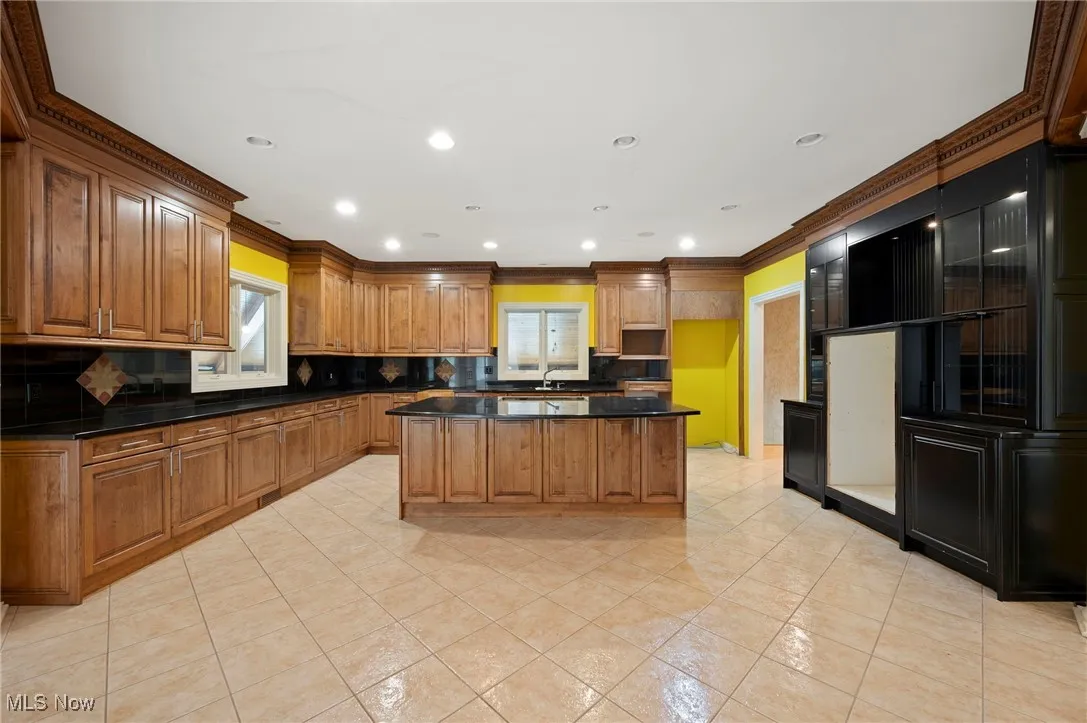 Kitchen featuring backsplash, dark countertops, light tile patterned flooring, crown molding, and brown cabinets