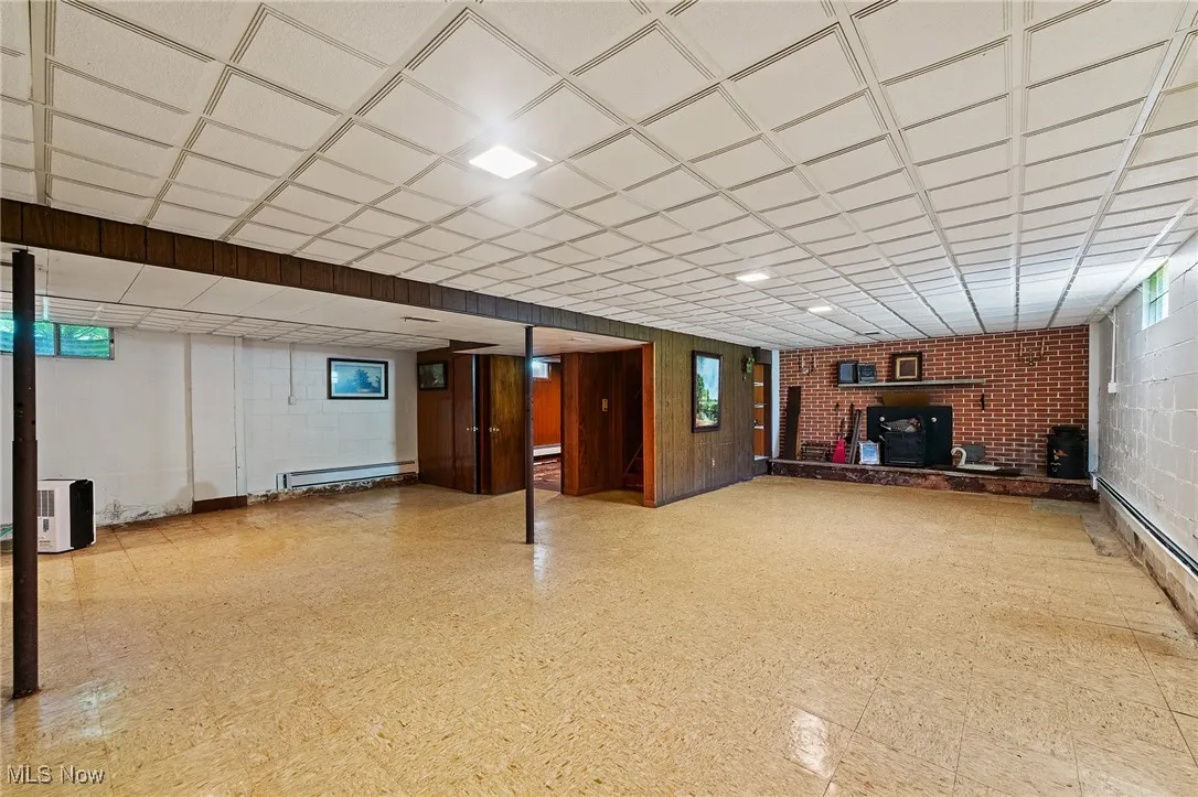Basement with a baseboard radiator, a paneled ceiling, and a wood stove