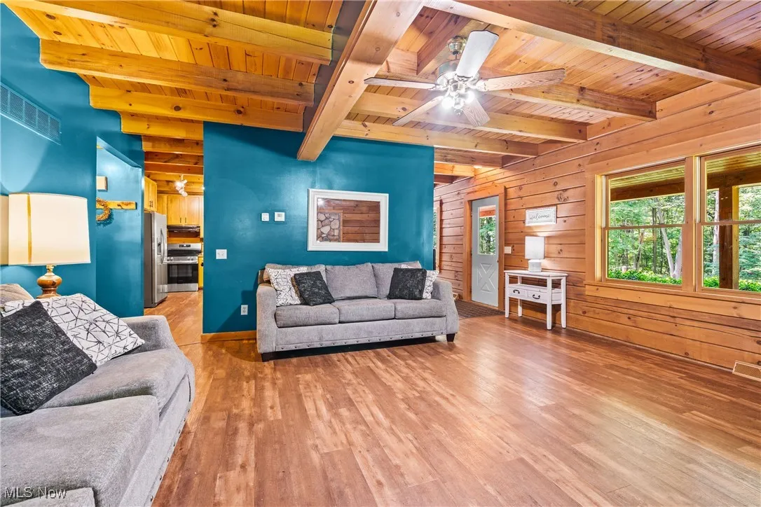 Living room with a wood ceiling with exposed beams, wood walls, wood-type flooring, and ceiling fan