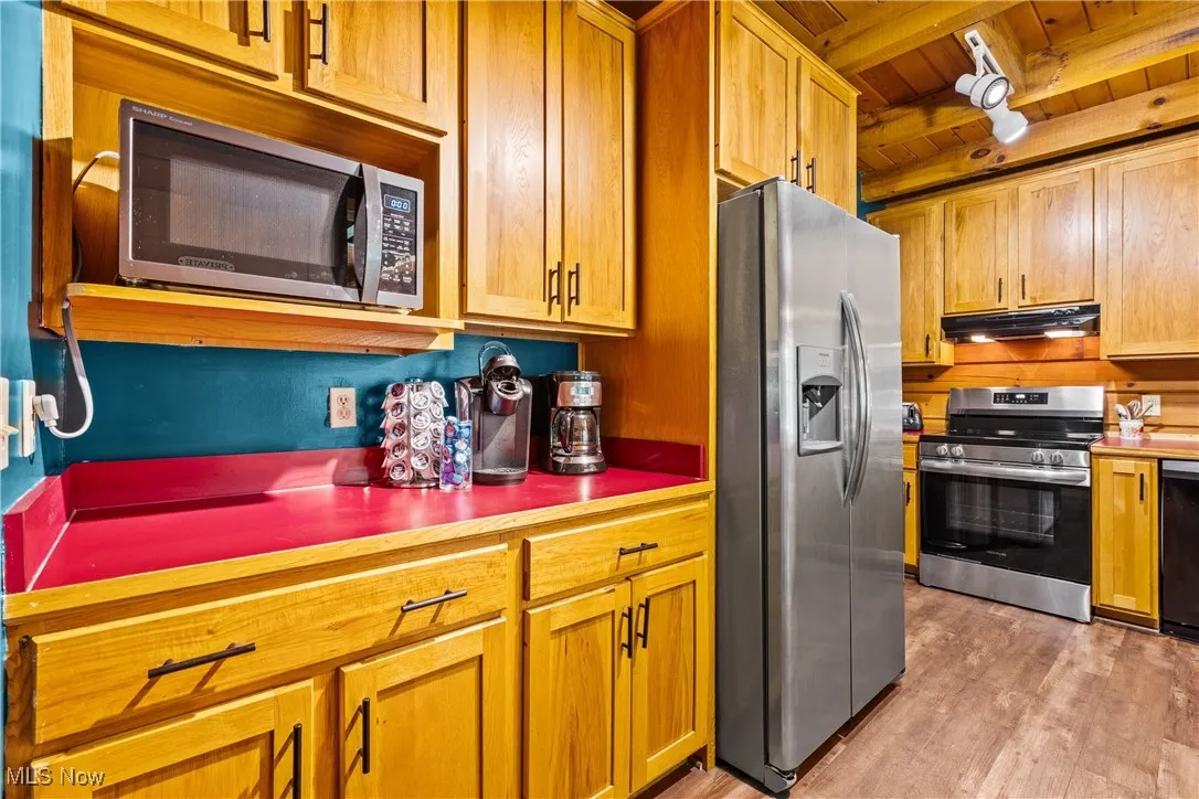 Kitchen featuring appliances with stainless steel finishes, under cabinet range hood, a wood ceiling with exposed beams, light wood finished floors, and rail lighting