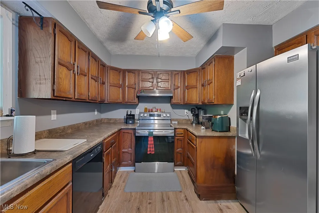 Kitchen featuring stainless steel appliances, under cabinet range hood, light wood-style flooring, brown cabinets, and a ceiling fan