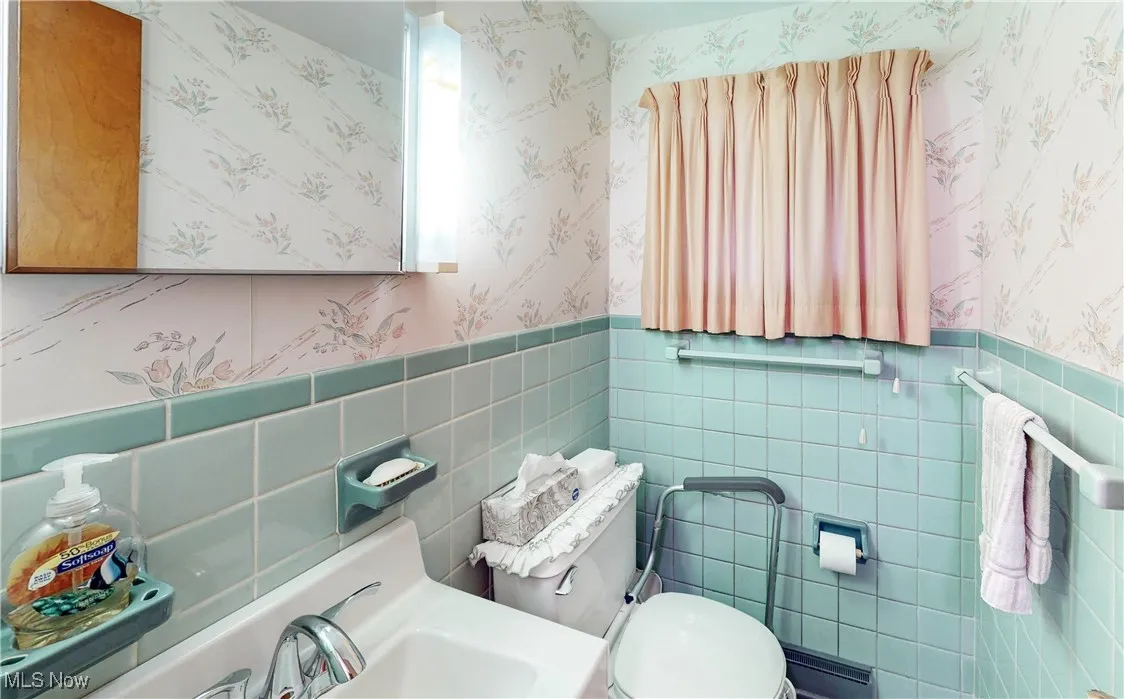 Half bathroom featuring wallpapered walls, tile walls, and wainscoting