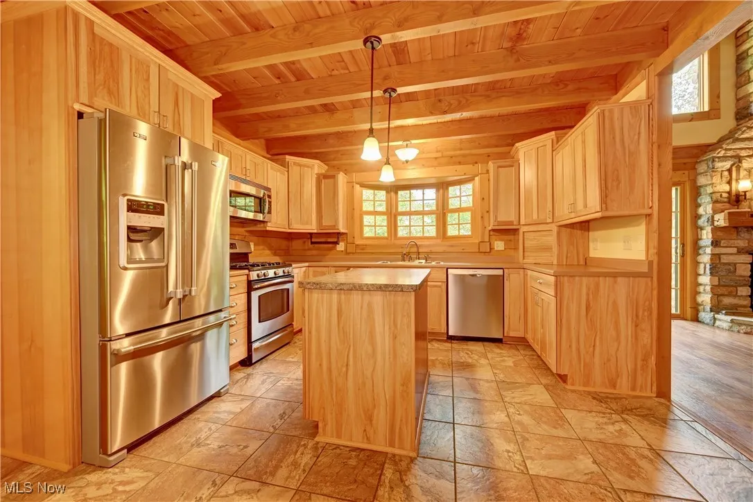 Kitchen featuring appliances with stainless steel finishes, a wood ceiling with exposed beams, light brown cabinetry, stone tile floors, and a kitchen island