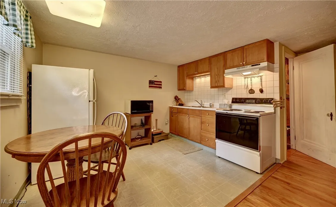 Kitchen featuring white appliances, under cabinet range hood, a textured ceiling, tasteful backsplash, and brown cabinetry